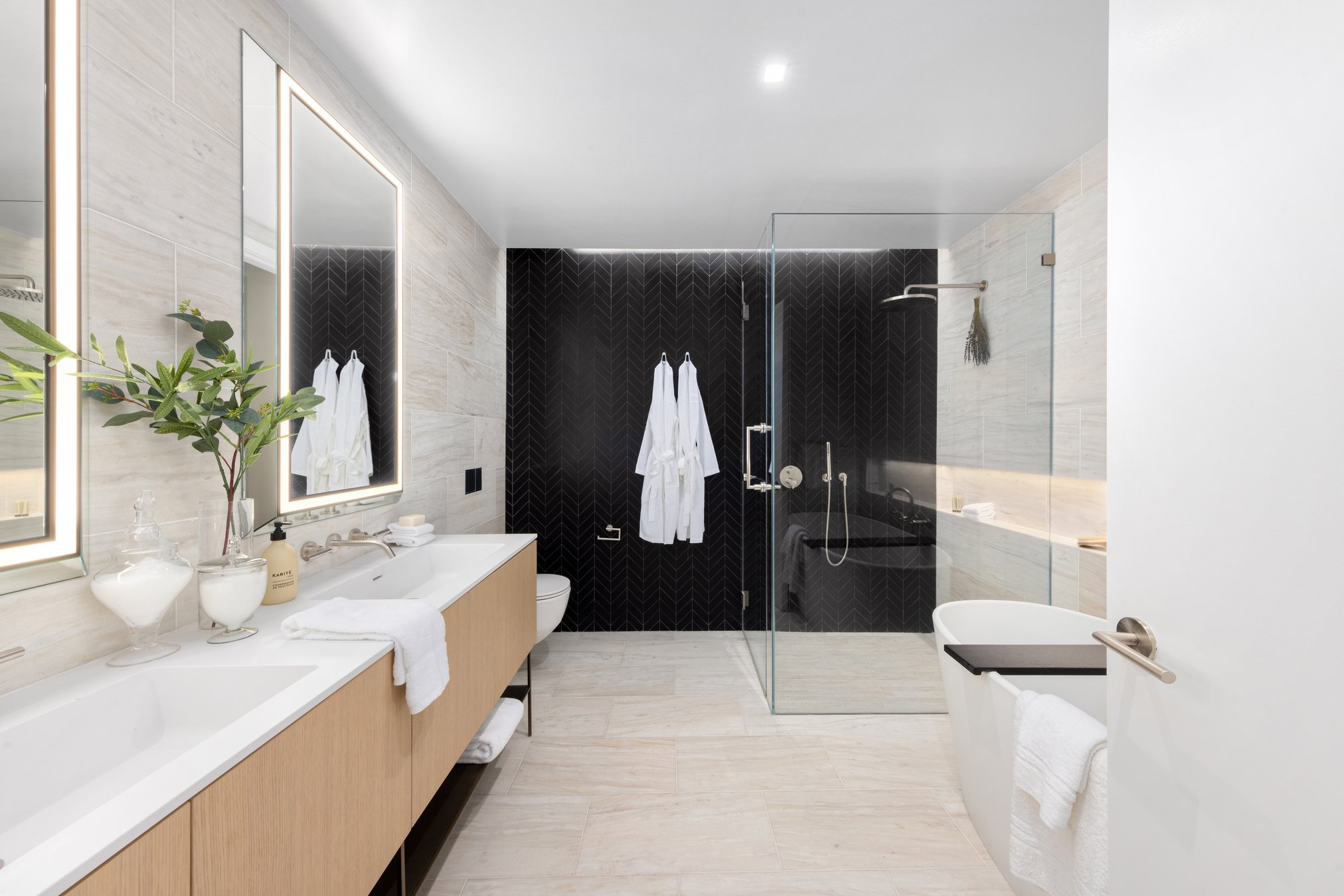 A bathroom with a glass-enclosed walk-in shower, hooks for towels or robes, toilet, a double sink and freestanding tub. The right wall has a full-length inset section cut into it that acts as lighting and a shelf