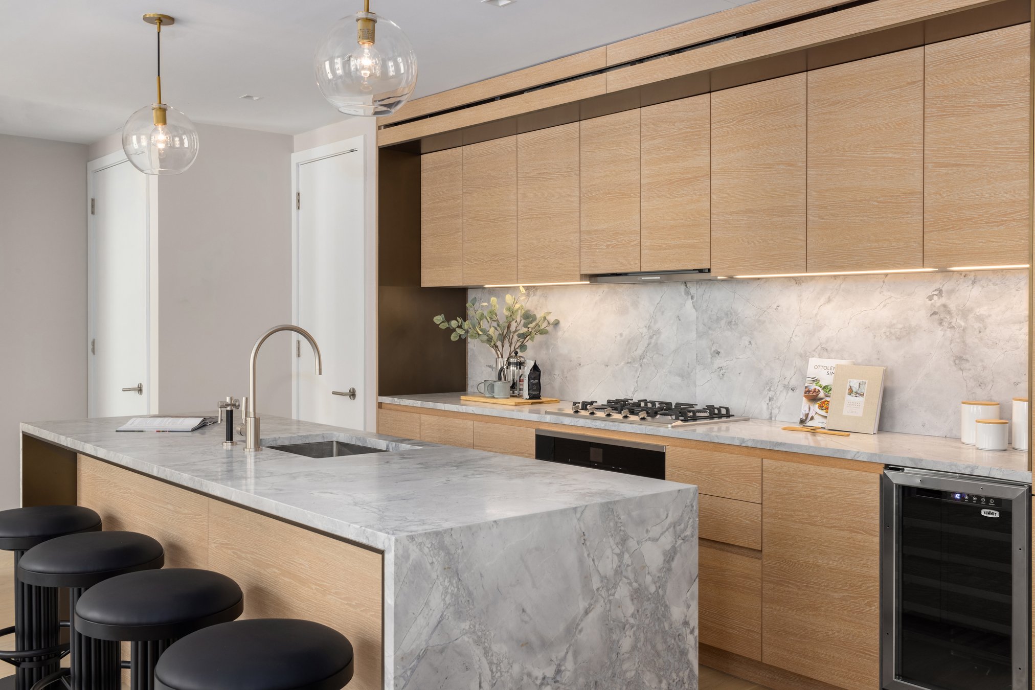 A render of the kitchen island with stools, a sink and some tomatoes in a basket, lights coming down from the ceiling above it, and the stove, cupboards and a wine rack behind it.