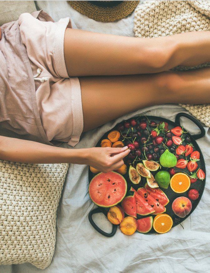 The waist to below the knees of a woman eating fruit on a bed