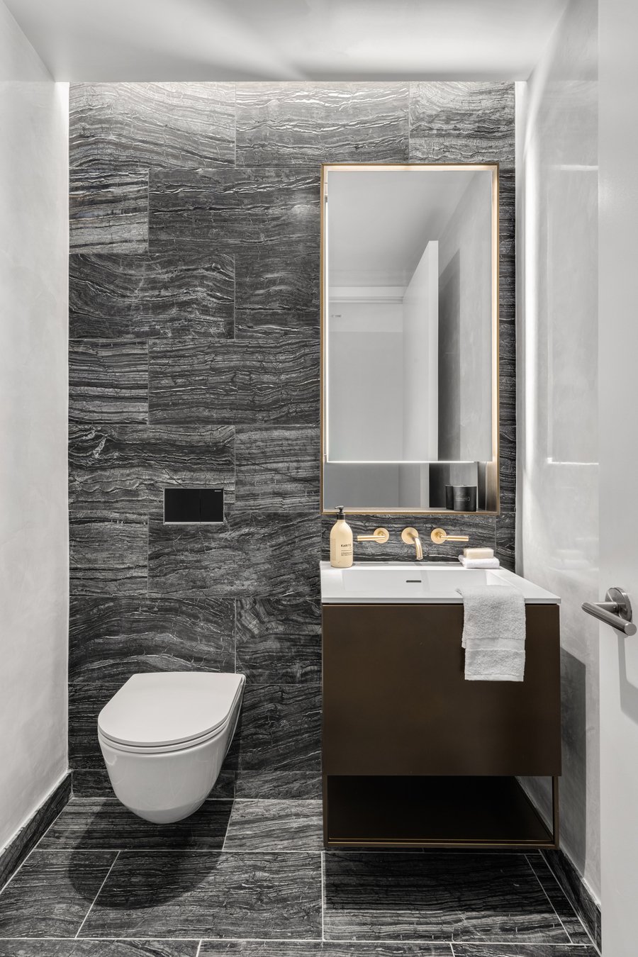 The powder room with a toilet mounted into the wall on the left and a sink with towels below it and a mirror above on the right. The sink has a vase with flowers and two tap handles surrounding a tap.
